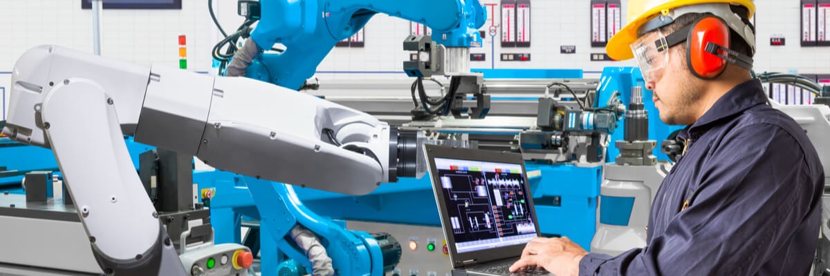 A worker on his laptop is standing next to manufacturing robots. 