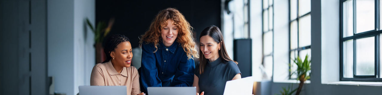 Diverse professionals collaborate on a laptop using Google Workspace in a sunlit, modern open-plan office.