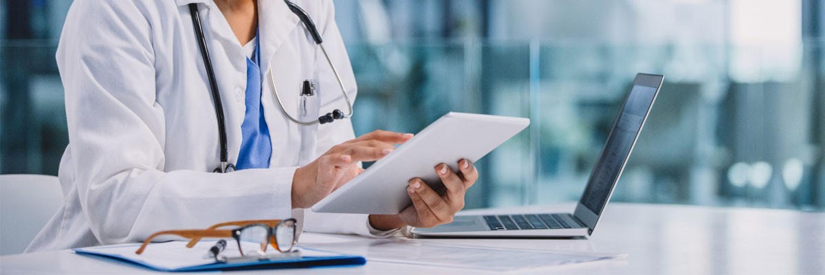 Doctor holding tablet device with patient data while sitting at desk with laptop computer