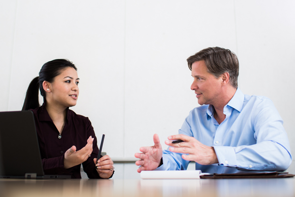 Man with pen and paper speaking to woman with laptop computer